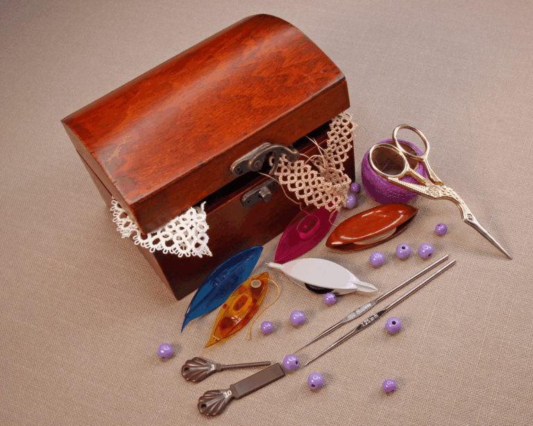 Open wooden box filled with shuttle tatting tools and accessories: colourful shuttles, small scissors, crochet hooks, beads, picot gauges, and delicate lace samples spilling over the edge.